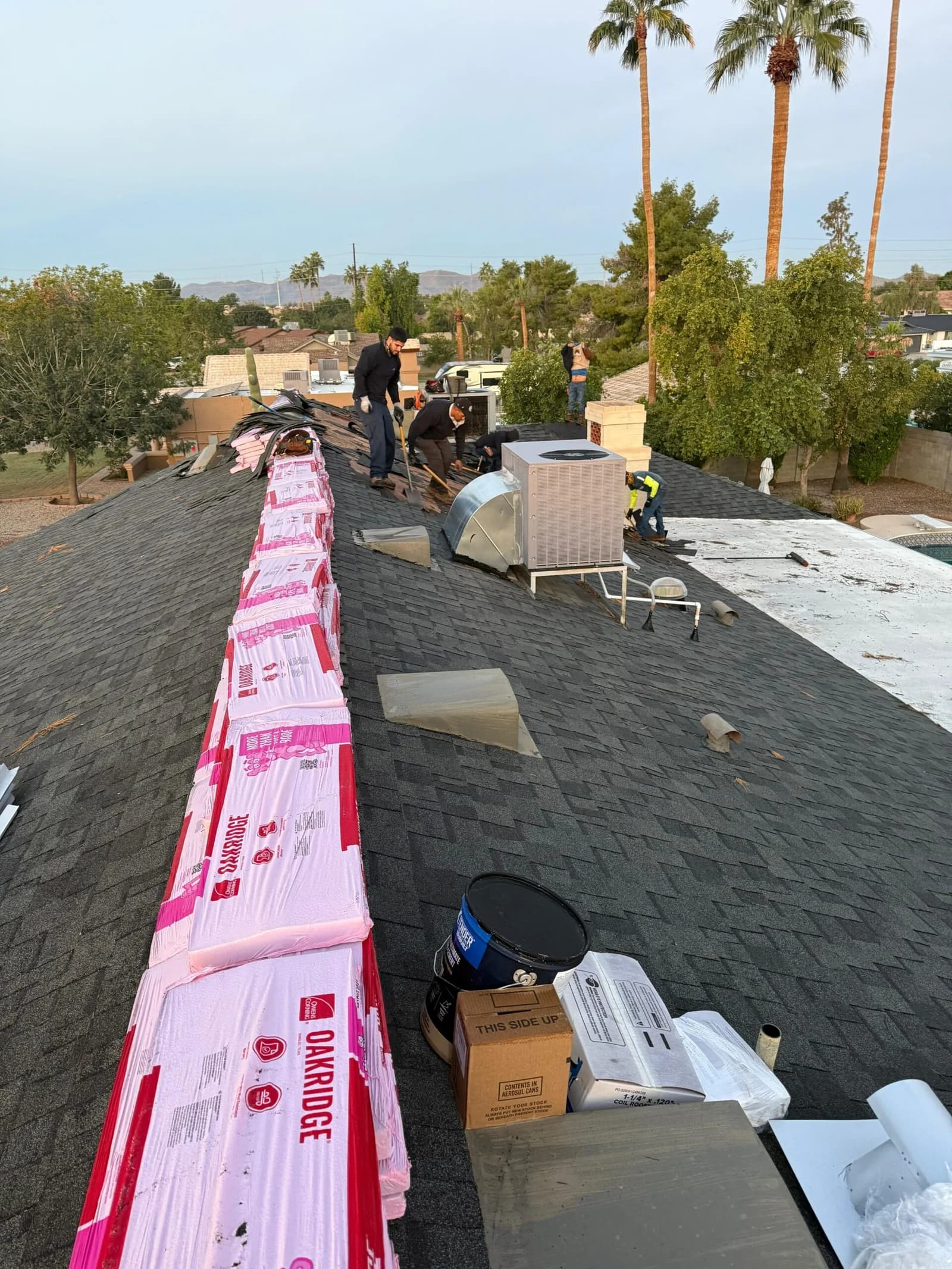 Roofers installing dark shingles on a residential roof with pink bundles along the peak.